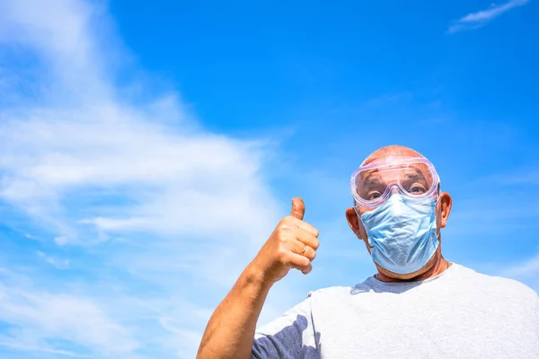 senior man wearing medical mask and goggles looking at the camera and shows thumbs up. Old man on a blue sky with clouds background