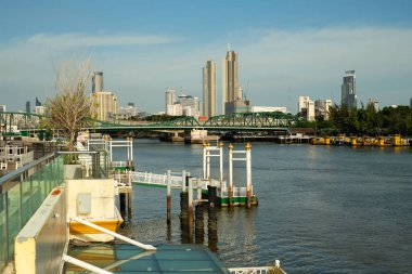 Köprüsü olan bir nehir kenarındaki beyaz rıhtımlar ve arka planı 2 olan binalar.