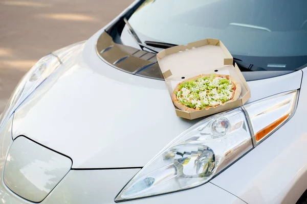 Pizza closeup on hood of the eco car. Safe delivery of pizza with green salad, tomatoes, cheese. Advertise in social network for restaurant delivery. Ecological delivering by electric car