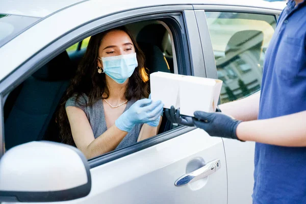 Young attractive girl in car gets 2 wok in box udon noodles with tempuru, shrimps, in hands and smiles. Courier in medical gloves. Udon noodles in white box delivery. Wok box udon advertise 1+1.