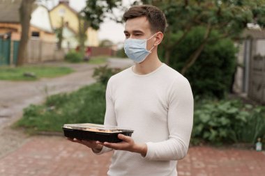 Suşi, arabayla çevrimiçi sağlıklı yiyecek dağıtım kutusuna koyuldu. Tıbbi maskeli, suşi kutulu bir kurye arabanın önünde duruyor. Japon mutfağı: ekmek, soya sosu, wasabi. Paket yemek için yönlendir