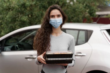 Suşi, arabayla sağlıklı yemek servisi kutusuna kondu. İki suşi kutusuyla tıbbi maskeli çekici bir kız arabanın önünde duruyor. Japon mutfağı: ekmek, soya sosu, wasabi. 1 + 1 restoran reklamı