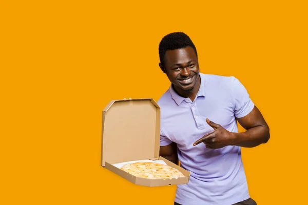 Tasty food pizza delivery from restaurant. Safety delivery. Pizza with cheese boards advertise. Isolated yellow background. Young african bearded courier points to pizza