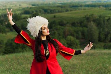 Georgian girl in white papakha dances national dance in red national dress on the green hills of Georgia background. Georgian culture lifestyle. clipart