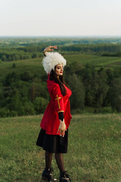 Georgian girl in white papakha and red national dress dances national dance named: rachuli, acharuli, osuri, shalaxo, mtiuluri. on the green hills of Georgia background. Georgian culture lifestyle.