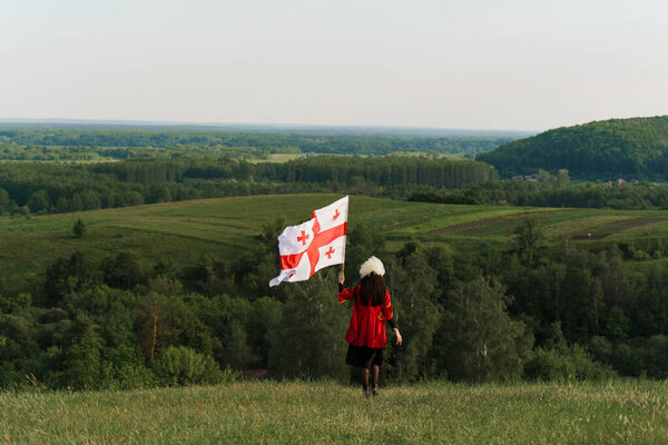 Georgian girl with national flag of Georgia in hands walks around green hills. Georgian culture lifestyle. Woman in papakha and red dress