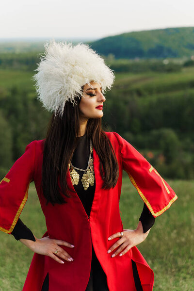 Georgian girl in papakha and red national dress with cross symbols. Attractive woman on the lake. Georgian culture lifestyle. Woman looks right side