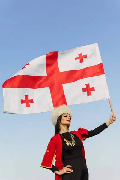 Georgian girl waving national flag of Georgia on blue sky background. Georgian culture lifestyle. Woman in papakha and red dress