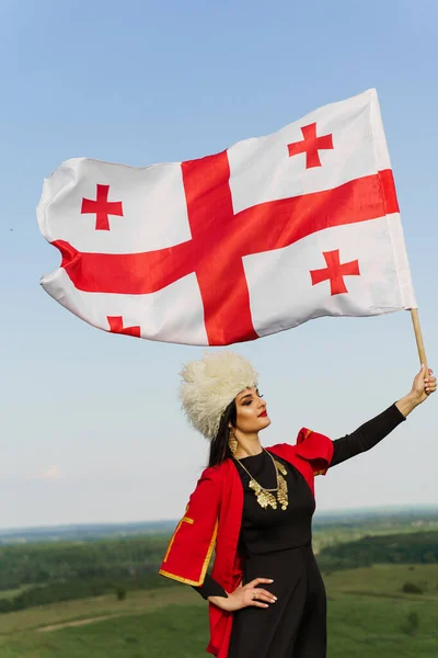 Georgian girl holds national flag of Georgia on blue sky background. Georgian culture lifestyle. Woman in papakha and red dress