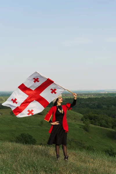 Georgian girl waving national flag of Georgia on blue sky background. Georgian culture lifestyle. Woman in papakha and red dress