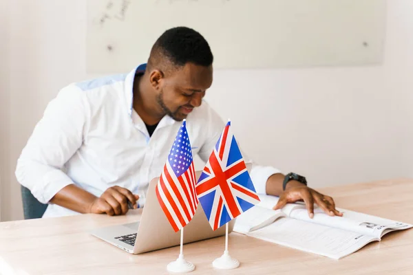 English and British flags in front. Black handsome translator uses his laptop for online work according social distancing. Scientist works at home on quarantine period.