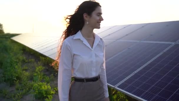 女性は日没時に太陽電池パネルの列の近くを歩き 笑顔で太陽電池を見ています 女性投資家は正式な白いシャツを着ている 家のための無料電気 地球の持続可能性 緑のエネルギー — ストック動画