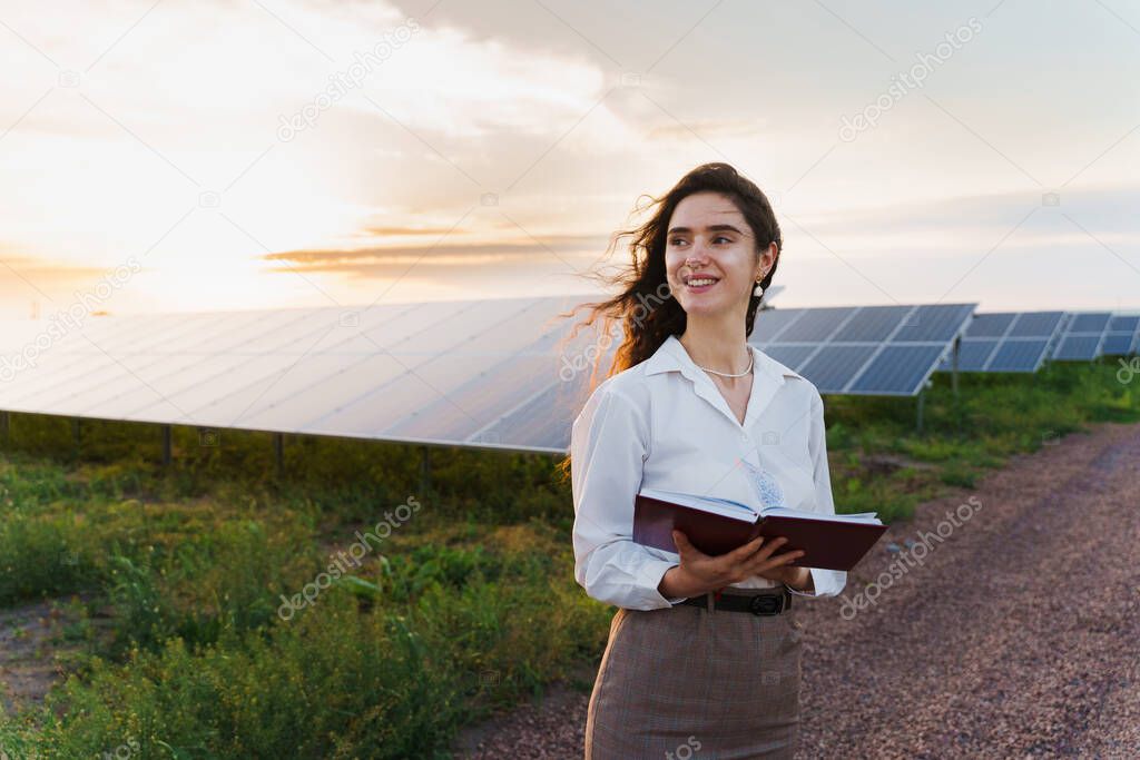 Investor woman stand and read book near solar panels row on the ground at sunset. Girl weared formal white shirt. Free electricity for home. Sustainability of planet. Green energy.