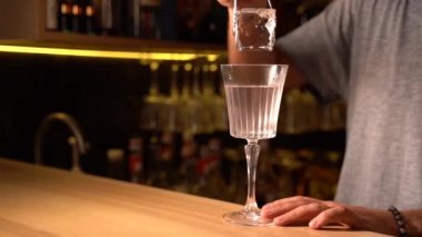Barmen cımbızla büyük bir buz parçası tutuyor ve Fransız 75 kokteylinin içine atıyor. Barthender barda alkol kokusu yapıyor.
