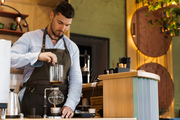 Handsome bearded barista mix freshly ground coffee in syphon device for coffee brewing in cafe. Syphon alternative method of making coffee. Scandinavian method of coffee making.