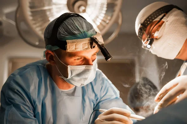 2 surgeons with headlamp do plastic operation in medical clinic. Chest augmentation plastic operation and correction in medical clinic. Group of professional medics in medical masks do plastic surgery.