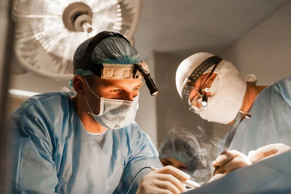 2 surgeons with headlamp do plastic operation in medical clinic. Chest augmentation plastic operation and correction in medical clinic. Group of professional medics in medical masks do plastic surgery.