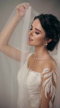 Gelinlik içindeki genç çekici gelin beyaz arka plan dikey videosundaki duvağa dokunuyor. Düğün ajansı ve gelinlik salonu için sosyal ağların adresi