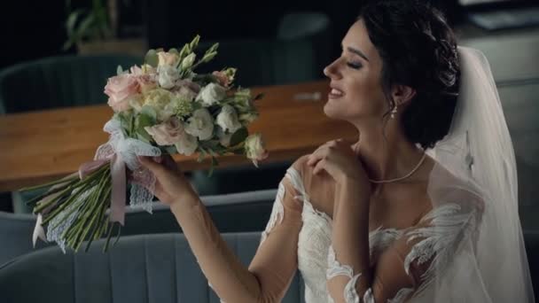 穿着婚纱婚纱的年轻漂亮的新娘看着花束笑着。在现代餐馆里穿婚纱的女孩。为婚介所的社交网络做广告. — 图库视频影像
