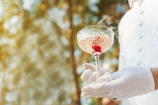 Waiter in white gloves gives wine glass with shampagne, red cherry, and white smoke of dry ice and gives to customer. Catering for wedding ceremony and business meeting. Empy left side for text.