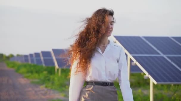 Mulher caminha perto de painéis solares remar no chão ao pôr do sol e sorrir e olhar para as células solares. Mulher investidora usa camisa branca formal. Eletricidade gratuita para casa. Sustentabilidade do planeta. Energia verde — Vídeo de Stock