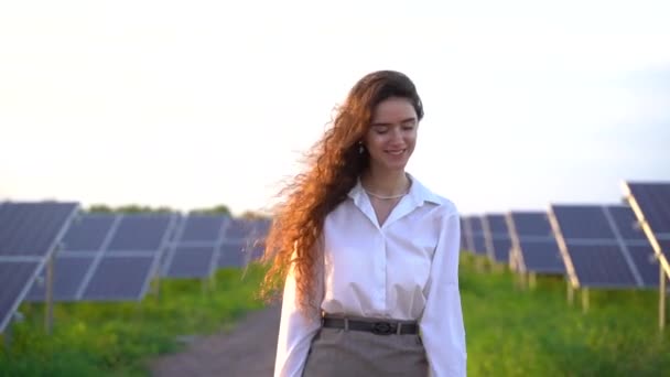 Γυναίκα περπατά κοντά σε ηλιακούς συλλέκτες σειρά στο έδαφος στο ηλιοβασίλεμα και χαμόγελο. Γυναίκα επενδυτής φοράει επίσημο λευκό πουκάμισο. Δωρεάν ηλεκτρικό ρεύμα για το σπίτι. Βιωσιμότητα του πλανήτη. Πράσινη ενέργεια — Αρχείο Βίντεο