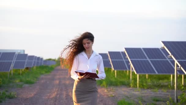 Kvinne går nær solpaneler ro på bakken ved solnedgang og smil og se på solceller. Investoren har på seg hvit skjorte. Gratis strøm hjemme. Planetens bærekraft. Grønn energi – stockvideo