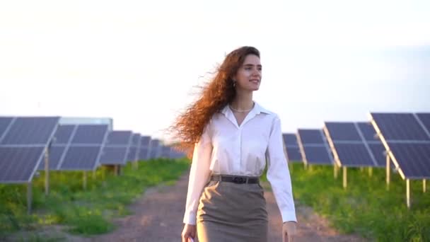 La mujer camina cerca de paneles solares reman en el suelo al atardecer y sonríen y miran las células solares. Inversionista mujer lleva camisa blanca formal. Electricidad gratuita para el hogar. Sostenibilidad del planeta. Energía verde — Vídeos de Stock