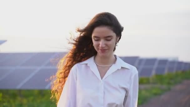 La donna cammina vicino ai pannelli solari fila a terra al tramonto e sorride e guarda le celle solari. Una donna investitrice indossa una camicia bianca formale. Elettricità gratuita per la casa. Sostenibilità del pianeta. Energia verde — Video Stock