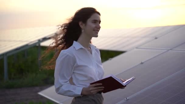Frau blickt auf Sonnenkollektoren, die bei Sonnenuntergang auf dem Boden liegen, lächelt und betrachtet Solarzellen. Investorin trägt formell weißes Hemd. Kostenloser Strom für zu Hause. Nachhaltigkeit des Planeten. Grüne Energie — Stockvideo
