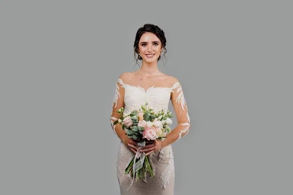 Bride with wedding bouquet smiles, looks in camera and touches her face. Attractive girl portrait for social networks. Girl in wedding gown on blank background
