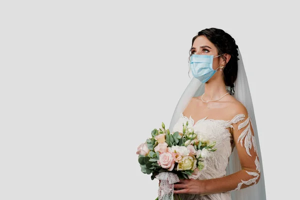 Bride in medical mask and wedding bouquet at coronavirus covid-19 quarantine period. Attractive girl in studio. Woman in medical mask looks left side with blank place for advert