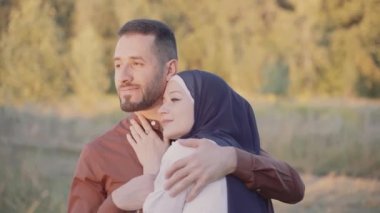 Sarılan, okşayan ve yeşil arka planda gün batımına bakan karışık çiftlerin mutlu Müslüman aşk hikayesi. Başörtülü kadın erkeğine bakıyor. Online randevu ajansına yönlendir