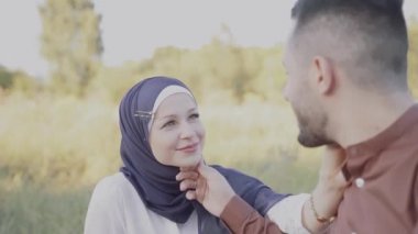 Müslüman aşk hikayesi videosu 4K 'da. Karışık çiftler sarılır, kucaklaşır ve gün batımında yeşil arka planda birbirlerine bakarlar. Başörtülü kadın erkeğine bakıyor. Online randevu ajansına yönlendir