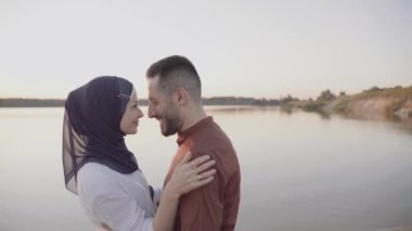 Müslüman aşk hikayesi videosu 4K 'da. Karışık bir çift kucaklaşır, okşar ve gölde gün batımını seyreder. Başörtülü kadın erkeğine bakıyor. Online randevu ajansına yönlendir