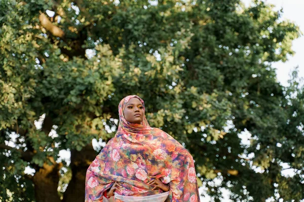 Attractive muslim black girl weared in hijab smiles and looks right side. African woman in national colorful dress on green background. Life style of black girl wearing in scarf named hijab