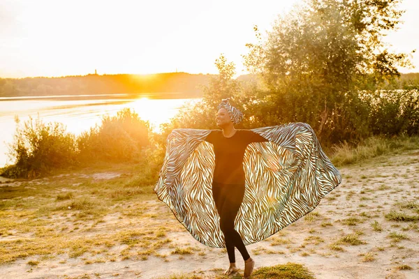 Happy muslim girl in trendy traditional clothes smiles and dances near the lake with sun light. Woman in traditional blue hijab. Cultures of the all of the world. African people lifestyle.