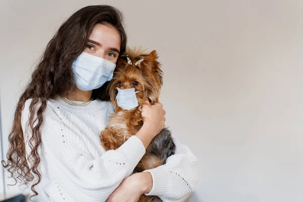 Köpeği Yorkshire Teriyeri olan çekici bir kadın. İkisi de beyaz arka planda koruyucu maske takıyor. Fotoğrafı kapat. Karantinaya alın. Önleyici önlemler. Sağlık ve hayvan bakımı. İnsanlar ve hayvanlar