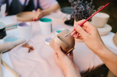 Zanaatkar seramik bardağı yumuşak mavi boyayla boyar, çömlek dekorasyonu ve el yapımı seramik tasarımın ayrıntılı sürecini yakalar. Kız masada mavi boyayla seramik bardak boyuyor.