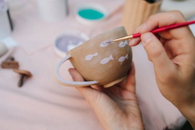 Fırça ve boya kullanarak seramik bardağı mor balıkla boyamak. Fırça darbeleriyle balık desenli kupa seramik resmi. Sanatçı, el yapımı kil bardağa narin mor balıklar çiziyor.