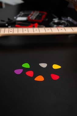 Gitar ve tamir aletleriyle siyah bir yüzey üzerinde renkli gitar penaları bulunuyor. Masanın üzerinde parlak renkli birden fazla gitar penası var. Gitar seçimleri ve arka planda araçlar