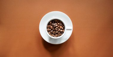 Kahverengi arka planda, beyaz kahve fincanında kavrulmuş kahve çekirdekleri. 