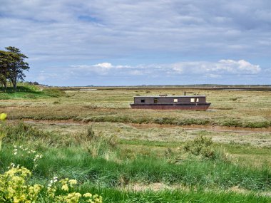 Brancaster Marsh 'taki bir teknenin profili. Kuzey Norfolk kıyısındaki Burnham Deepdale yakınlarında. Ufukta yürüyen yürüyüşçüler ve ufukta Royal West Norfolk Golf Kulübü var..
