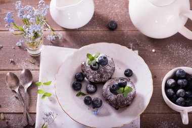 Çikolatalı kekler (kapkekler) masanın üzerinde. Kekler üzerine yaban mersini ve yeşil yaprak süslemeli nane. Beyaz tabaklarla servis edilen güzel ve zarif.