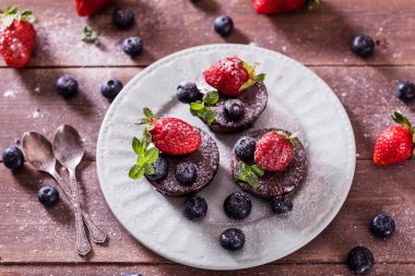 Çikolatalı kekler (kapkekler) masanın üzerinde. Kekler üzerine yaban mersini, çilek ve yeşil yaprak süslemesi. Beyaz tabaklarla servis edilen güzel ve zarif.