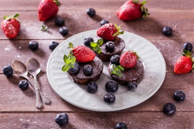 Çikolatalı kekler (kapkekler) masanın üzerinde. Kekler üzerine yaban mersini, çilek ve yeşil yaprak süslemesi. Beyaz tabaklarla servis edilen güzel ve zarif.