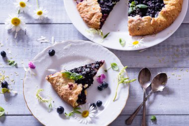 Ev yapımı çıtır yaban mersinli turta veya galette ve nane. Yaz, parlak, lezzetli turtalar çiçeklerle çerçevelenmiş ve böğürtlenli otlar
