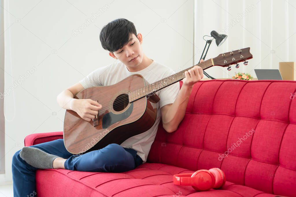 A young Asian man is at home happily. He is unemployed He is playing guitar and singing to relax.