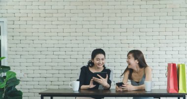 Gülen ve çekici iki Asyalı kadın bir kafede kahve hakkında konuşan arkadaşlar. Güzel ve kendine güvenen kadınlar internetten alışveriş yapmak için tablet kullanıyorlar..
