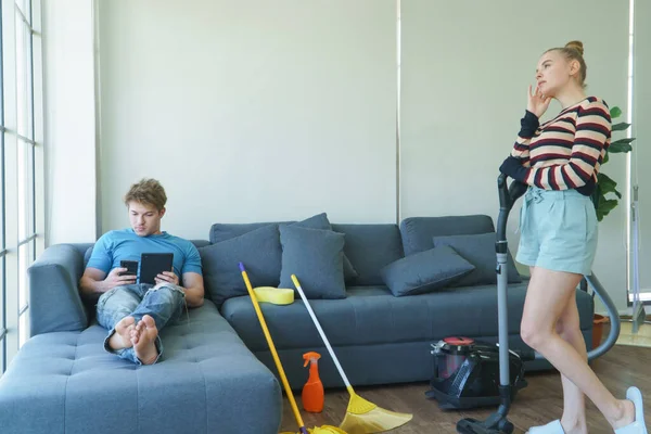 A young couple of Caucasian people do chores together. Beautiful women are cleaning the floor using vacuum cleaners. Handsome men are lazy to sleep and use tablets on the sofa.